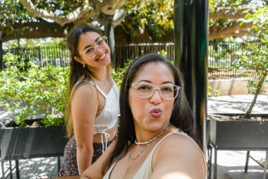 Selfie portrait of two Venezuelan Latina young women making funny faces, smiling and enjoying, taking photos with the phone, sitting in a restaurant, phone perspective