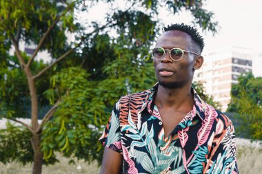 portrait of young man of african ethnicity, wearing glasses and colorful clothes, standing in a public square looking at the camera outdoors, with trees in the background and copy space.