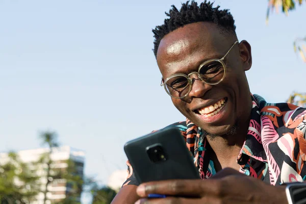 happy to earn money from digital investments, portrait of young man of African ethnicity, sitting outdoors smiling watching phone, business and technology concept. copy space.