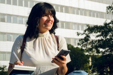 Venezuela kökenli genç Latin bir bayan öğrenci, kısa saç ve günlük kıyafetler, gülümseyen ve düşünceli bir şekilde üniversitenin dışında oturuyor telefonunu kullanıyor ve defterine notlar yazıyor..