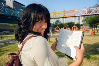 Genç Venezuelalı Latin bir kadın, üniversite öğrencisi, açık bir kitap gösteriyor, kameraya bakıyor ve gülümsüyor..