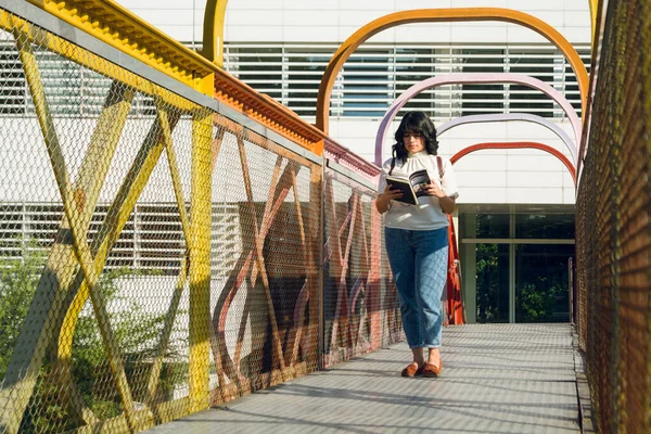young college student woman, walking on footbridge in college, reading book, school concept, copy space.