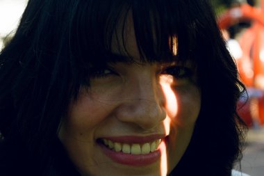 Genç Latin kadının yakın plan portresi. Yüzünde bir ışık çizgisi ile gölgede gülümsüyor ve kameraya bakıyor..