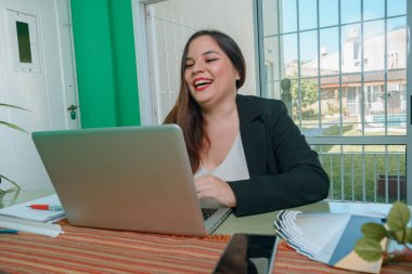 Evde dizüstü bilgisayarı üzerinde çalışırken yüksek sesle gülümseyen, evinde oturup dizüstü bilgisayara bakan aşırı neşeli genç bir kadın. Girişimci insanlar konsepti, alanı kopyala.