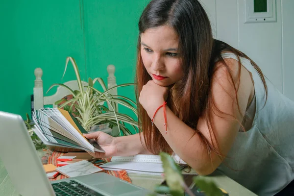 closeup of young hispanic latina woman, interior designer with wooden samples in her hand thinking which one to use for her work, leaning on the table looking at the laptop, with one hand on the chin.
