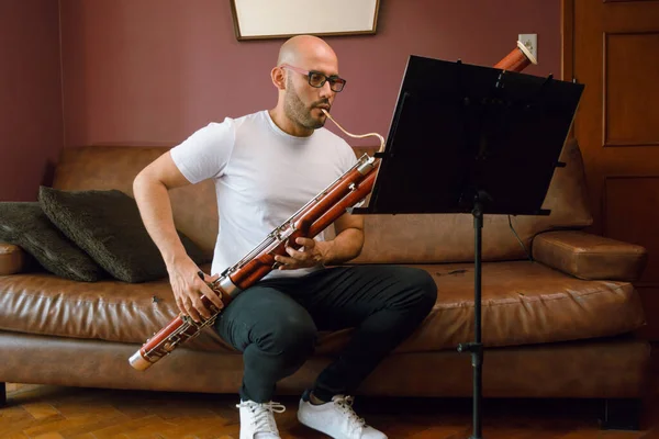 stock image young latino musician man, bald with glasses and white t-shirt, at home studying classical music playing the bassoon sitting on the sofa in the living room, reading sheet music