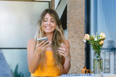 Sarı elbiseli genç Latin bir kadın evinin balkonunda oturuyor. Gülümsüyor ve telefonunu kullanıyor. Masasında çiçekler, insanlar ve teknoloji konsepti var..