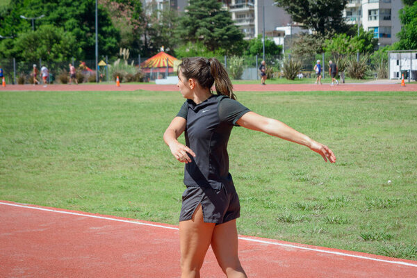 young argentinian latin woman, stretching arms and shoulders on public running track in park for training, wears black sportswear.