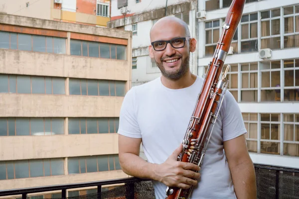 stock image Waist-up portrait of young orchestra musician Venezuelan Latin man with beard, glasses and white clothes, standing smiling holding a bassoon on the balcony of his apartment, looking at the camera.