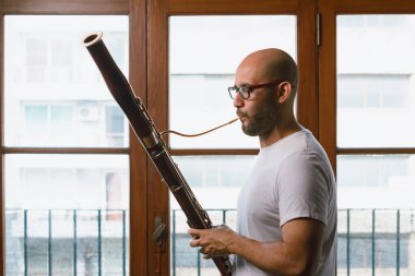 Orta boy, sakallı, gözlüklü ve tırnaklı genç Venezuelalı bir adamın fagot çalarken, arka planda pencereleri olan fotoğrafı..
