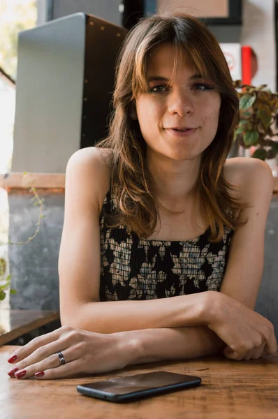 vertical portrait of young transgender latina woman sitting in a restaurant leaning on the table, calmly looking at the camera waiting for the waiter, with the phone on the table.