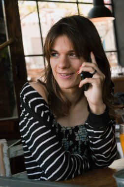 Genç transseksüel Latin kadının dikey portresi. Telefonda konuşuyor. Restoranda pencereden dışarı bakıyor..