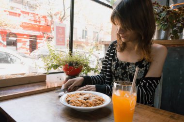 Arjantinli etnik kökenli genç transseksüel Latin bir kadın, bir restoranda çatal bıçakla portakal suyuyla bir tabak erişte yemeye başlıyor. yiyecek ve içecek konsepti, kopyalama alanı.