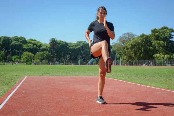 young latina woman of argentinian ethnicity in black sportswear starting warm up exercises on the running track with one leg raised and slightly bent to the front