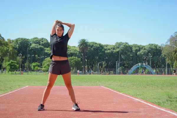 young latin woman of argentinian ethnicity in black sportswear, standing outdoors stretching arms and shoulders before training, looking at camera, copy space