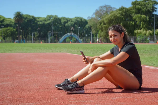 年轻的阿根提尼拉丁女人穿着黑色运动服 坐在跑道上 用手机微笑着看着相机 技术概念 复制空间 — 图库照片