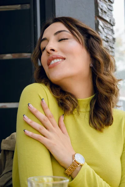 Stock image beautiful young hispanic latina woman of argentinian ethnicity with a green sweater, brown wavy hair, sitting in front of the cafeteria posing sensually with her eyes closed.