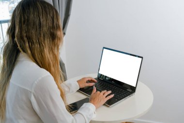 Tanımlanamayan genç bir kadının arka görüntüsü. Oturma odasında, boş ekranlı dizüstü bilgisayar kullanarak oturuyor. Fotokopi odası ve arka planında beyaz duvar var..