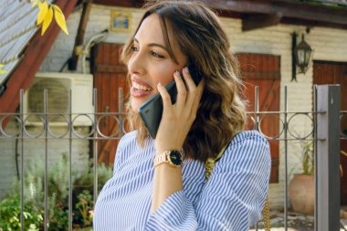 Mutlu Arjantinli etnik kadın evinin önünde telefonla konuşan, gülümseyen ve önüne bakan, zarif kıyafetler giyen, kahverengi, gevşek saçlı, kopyalama alanı olan.