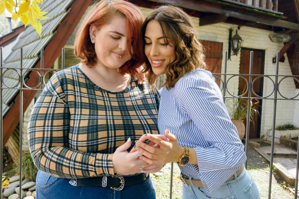 İki Arjantinli Latin bayan arkadaş, günlük giysiler içinde mutlu bir şekilde gülümsüyorlar, evin dışında durup telefona bakıyorlar, mesajları, bildirileri ve sosyal ağları kontrol ediyorlar.