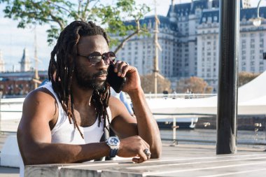 Rastalı, sakallı, güneş gözlüklü, beyaz tişörtlü, gün batımında dışarıda oturan Puerto Madero Arjantin 'de telefonla konuşan genç siyahi bir adam..