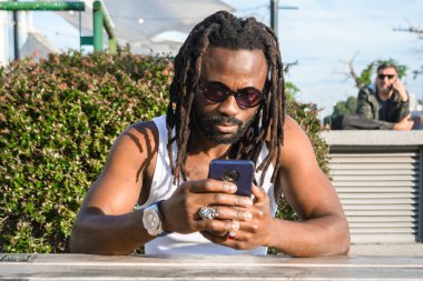 Afrika kökenli genç siyahi bir adamın ön görüntüsü. Rastalı ve güneş gözlüklü. Dışarıda oturmuş telefonuna dikkatle bakıyor..