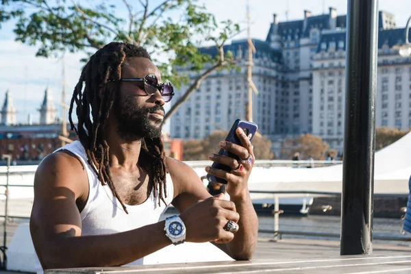 Rastalı, sakallı, güneş gözlüklü, beyaz tişörtlü, şüpheli bir yüzle dışarıda oturan, düşünen, telefonuna bakan, sosyal ağların dikkatini dağıttığı internette gezinen siyah bir adam..