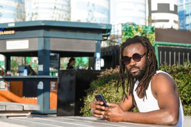 Afrika kökenli genç bir adamın portresi. Rastalı ve güneş gözlüklü. Açık hava yemek fuarında oturmuş garson tarafından servis edilmeyi bekliyor..