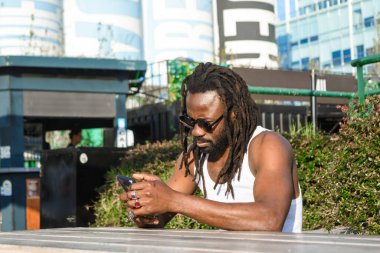 Afrika kökenli genç siyahi adam, açık hava yemek fuarında oturuyor, telefon kullanıyor, sosyal medya ile eğleniyor, yemeğini, yaşam tarzını, kopyalama alanını bekliyor..