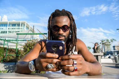 Saçları rastalı ve Afrika kökenli güneş gözlüklü genç siyahi adamın ön görüntüsü. Açık havada oturuyor, telefon kullanıyor, internette sörf yapıyor, sosyal medyayı kontrol ediyor..