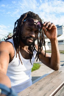 Rastalı ve sakallı genç Afrikalı bir adamın dikey selfie portresi, gün batımında dışarıda oturuyor, kızgın bir yüzle kameraya bakıyor, telefonla fotoğraf çekiyor.