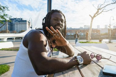 Rastalı ve sakallı genç bir turistin portresi. Dışarıda oturmuş kameraya el sallıyor ve gülümsüyor. Gün batımında bir masada oturuyor..
