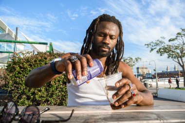 Rastalı genç siyahi adamın ön görüntüsü, gün batımında dışarıda oturuyor, plastik bir bardakta içecek ve kendini tazelemek için soda servisi yapıyor.