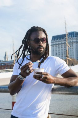 Afrika kökenli, rastalı, sakallı ve güneş gözlüklü genç bir adam. Buenos Aires 'te gün batımının tadını çıkarıyor. Puerto Madero' daki hendeğin yanındaki parmaklıklara yaslanıyor.