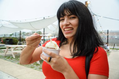 Genç Venezuelalı Latin kadın, plastik kaşıkla küçük bir fincan vanilyalı ve çikolatalı dondurma yerken kameraya bakıp mutlu bir şekilde gülümsüyor.