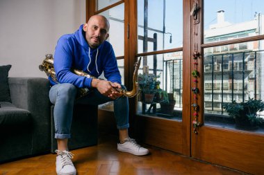 Venezüella kökenli genç Latin bir adamın portresi. Saksafonuyla evde, koltukta oturmuş kameraya bakıyor..