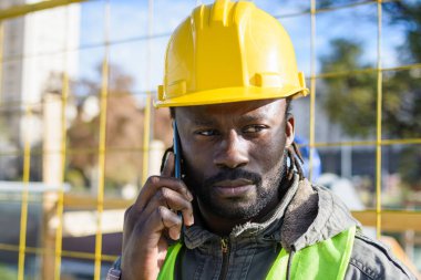 Sarı güvenlik kaskı takan, açık havada duran, telefon konuşmasında talimatları dikkatle dinleyen genç, siyah bir inşaat işçisi. Boşluğu kopyala.