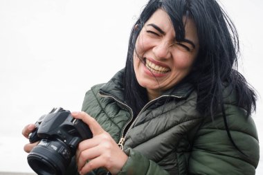 Venezuela etnik kökenli genç bir kadın, dijital kamerasıyla mutlu bir şekilde gülümsüyor soğuk ve bulutlu bir günde sosyal ağları için içerik yaratıyor..