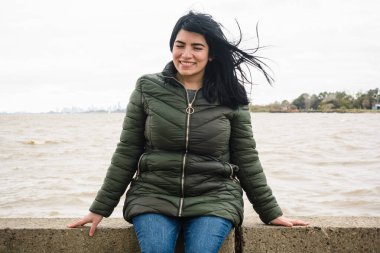 Genç Latin Venezuelalı turist kadın, gülümseyen ve kameraya bakan, arka planda nehir ve gökyüzü, turizm konsepti, kopyalama alanı olan bir iskelede oturuyor..