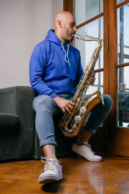 Venezuelalı genç bir adamın dikey portresi. Evdeki kanepede oturmuş saksafon çalıyor.