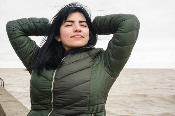 young latina tourist venezuelan woman with her eyes closed and her hands behind her head, is resting calm and serene, breathing and feeling the air on her face, on the pier next to the river