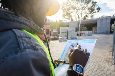 Afrika kökenli, sarı başlık ve yelek giyen siyah mühendis dikiz aynasından, klasördeki inşaat alanı malzemeleriyle not alarak, arka plan malzemeleriyle alanı kopyalayarak,.