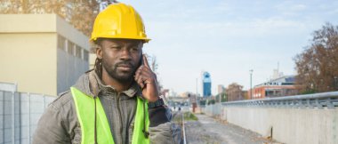 Afrika kökenli, sarı miğferli ve yansıtıcı yelekli müteahhit tren raylarında açık havada telefonla konuşuyor..