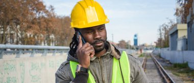 Afrika kökenli, sarı miğferli ve yansıtıcı yelekli müteahhit tren raylarında açık havada telefonla konuşuyor..