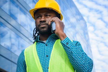 Sarı güvenlik kaskı ve yansıtıcı yelek giyen siyah erkek mühendisin portresi, açık havada telefonla konuşuyor ve etrafa bakıyor, arka planda ticari binalar ve uzay kopyaları var..
