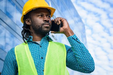 Sarı güvenlik kaskı ve yansıtıcı yelek giyen siyah erkek mühendisin portresi, açık havada telefonla konuşuyor ve etrafa bakıyor, arka planda ticari binalar ve uzay kopyaları var..