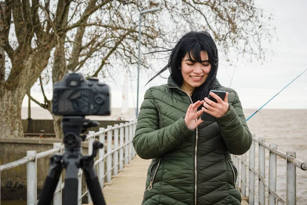 Genç Latin kadın gezgin içerik yaratıcısı gülümsüyor açık havada fotoğraf ve video çekiyor, dijital kamerasını telefonundaki bir uygulama ile kontrol ediyor, teknoloji kavramı, kopyalama alanı.