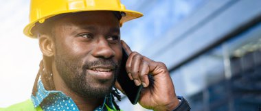 Sakallı, rastalı, sarı kasklı siyah mühendisin yakın plan pankartı. Telefonda konuşuyor, gülümsüyor. İyi haberleri duyuyor ve etrafa bakıyor..