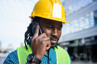 Afrikalı etnik kökenli yetişkin mühendis dışarıda güvenlik kaskıyla duruyor, iş sırasında telefonla konuşuyor, telefonla görüşüyor ve dikkatle dinliyor..