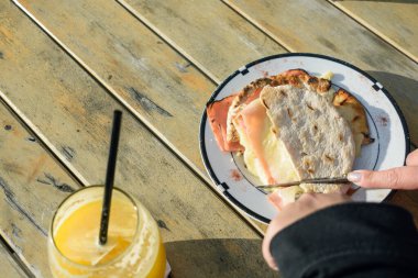 Gün batımında restoranın dışındaki ahşap masada çatal ve bıçakla doğranmış jambonlu ve peynirli sandviç dilimlenmiş beyaz kadın elleri, fotokopi alanı, yemek konsepti.